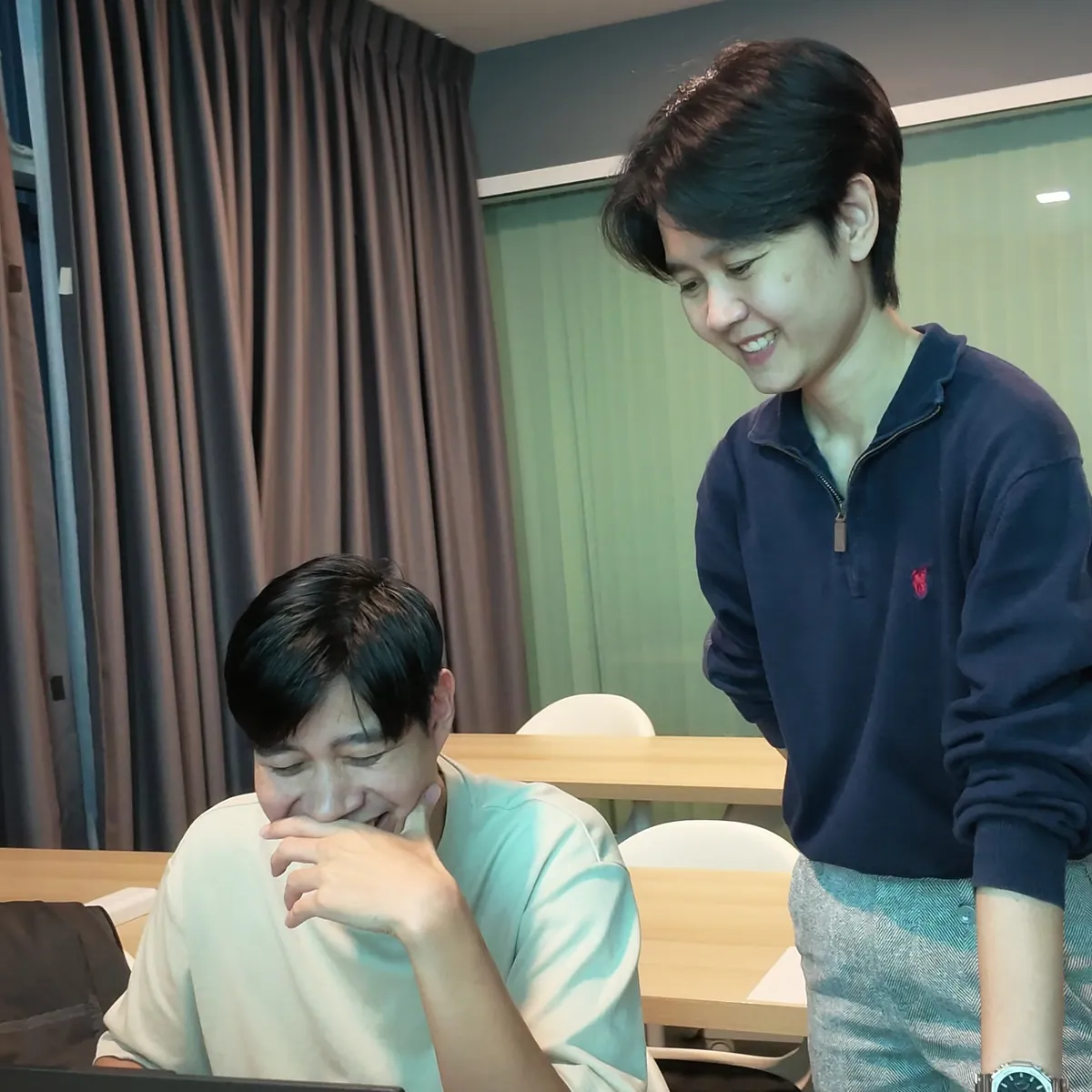 ผู้เรียนหัวเราะ ผู้ช่วยอาจารย์ยืนยิ้มข้างโต๊ะ